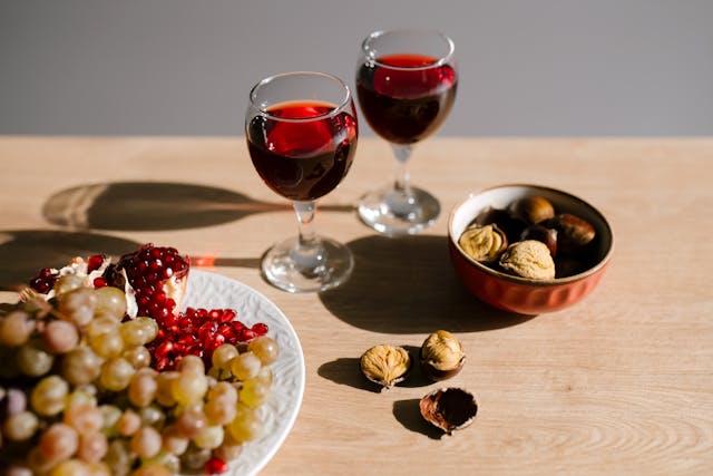 Mesa con dos copas de vino tinto, un plato de fruta, uvas y granadas al lado de un cuenco con castañas asadas abiertas