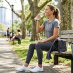 Mujer tomando agua en un parque sentada en un banco público, después de hacer ejercicio
