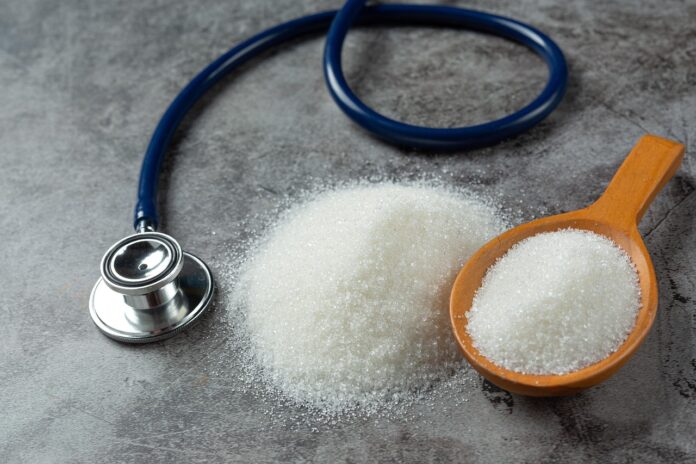 Estetoscopio-azucar-control-diabetes Una mesa con una cuchara que contiene azñucar blanca y al lado hay un estetoscopio