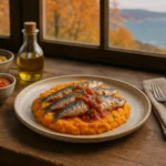 Un plato con sardinas sobre una base de puré de verduras o boniato. Al lado de una ventana, y al fondo se ve el mar y un paisaje otoñal