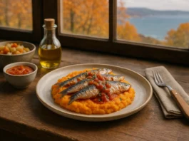 Un plato con sardinas sobre una base de puré de verduras o boniato. Al lado de una ventana, y al fondo se ve el mar y un paisaje otoñal