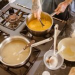 Una cocina, donde se intuye la mano del cocinero. Tiene al fuego hasta 3 calderos donde se está cocinando varios caldos