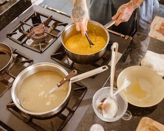 Una cocina, donde se intuye la mano del cocinero. Tiene al fuego hasta 3 calderos donde se está cocinando varios caldos