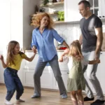 Día del Padre con diabetes: ideas para celebrar en familia Familia con dos niñas bailando y divirtiéndose en la cocina de casa, mostrando un momento cotidiano de actividad física y bienestar en familia.
