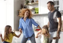 Familia con dos niñas bailando y divirtiéndose en la cocina de casa, mostrando un momento cotidiano de actividad física y bienestar en familia.