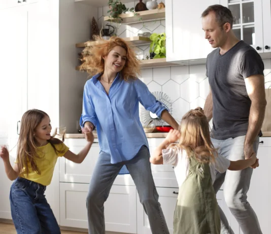 Familia con dos niñas bailando y divirtiéndose en la cocina de casa, mostrando un momento cotidiano de actividad física y bienestar en familia.