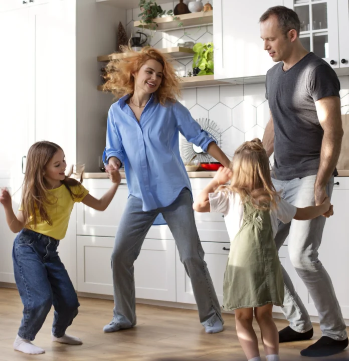 Familia con dos niñas bailando y divirtiéndose en la cocina de casa, mostrando un momento cotidiano de actividad física y bienestar en familia.