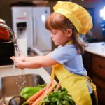Una niña con delantal amarillo delante de un fregadero, lista parece para ponerse a cocinar